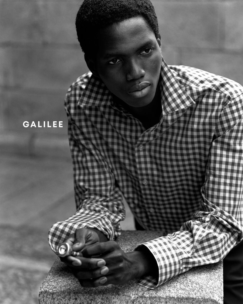 Black and white portrait of a Black male model in a checkered button-down shirt, leaning against a stone wall and holding an object.