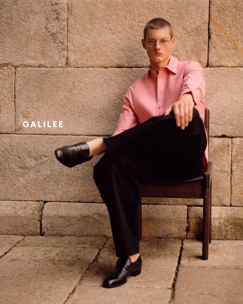 Male fashion model poses outdoors sitting on a chair in a pink shirt and black pants against a stone wall background.