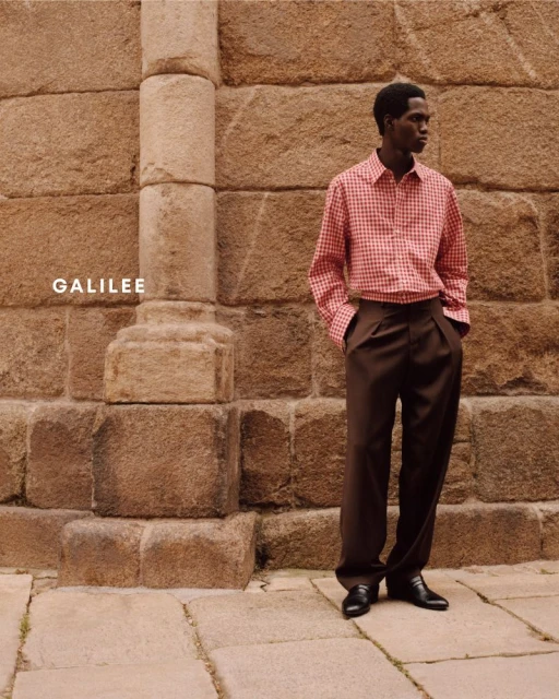 A dark-skinned man poses in a red and white checkered shirt and brown slacks in front of a tan stone wall.