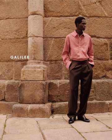 A dark-skinned man poses in a red and white checkered shirt and brown slacks in front of a tan stone wall.