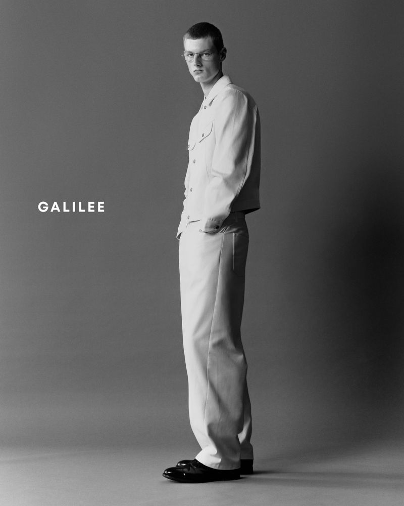 Full-length studio shot of an adult male model in matching cream-colored jacket and trousers, with black shoes, against a grey background.