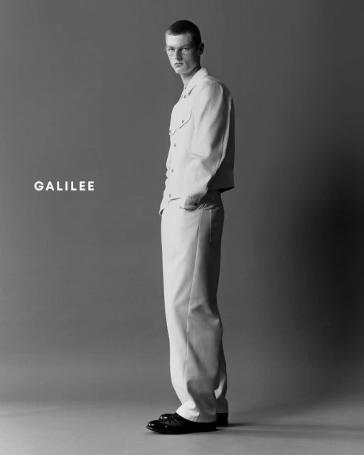Full-length studio shot of an adult male model in matching cream-colored jacket and trousers, with black shoes, against a grey background.