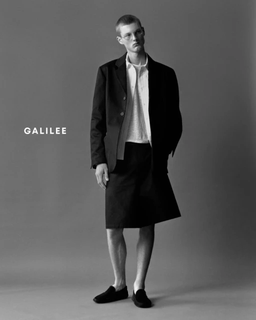 Black-and-white studio shot features a slender adult male wearing a black blazer and skirt, gazing slightly upward with a serious expression.