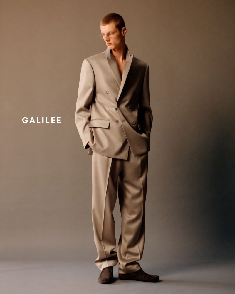 Studio shot of a young adult male model posing in a tailored double-breasted suit and dress shoes against a neutral grey background.