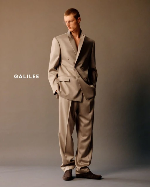 Studio shot of a young adult male model posing in a tailored double-breasted suit and dress shoes against a neutral grey background.