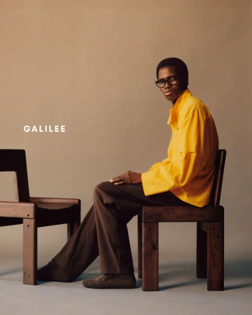 A person seated on a dark wood chair wearing glasses, a yellow shirt, and brown pants next to another dark wood chair in studio.