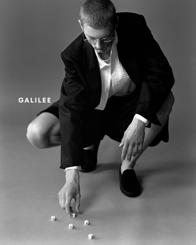Monochrome portrait of a person in a suit jacket and shorts, squatting, and arranging dice on the floor in a studio setting.