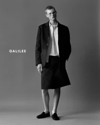 Man wearing a black blazer and shorts with glasses standing against a plain grey studio backdrop