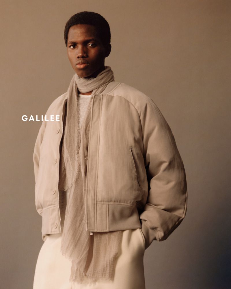 Black man wearing a beige bomber jacket, matching scarf, and light trousers against a neutral studio backdrop.