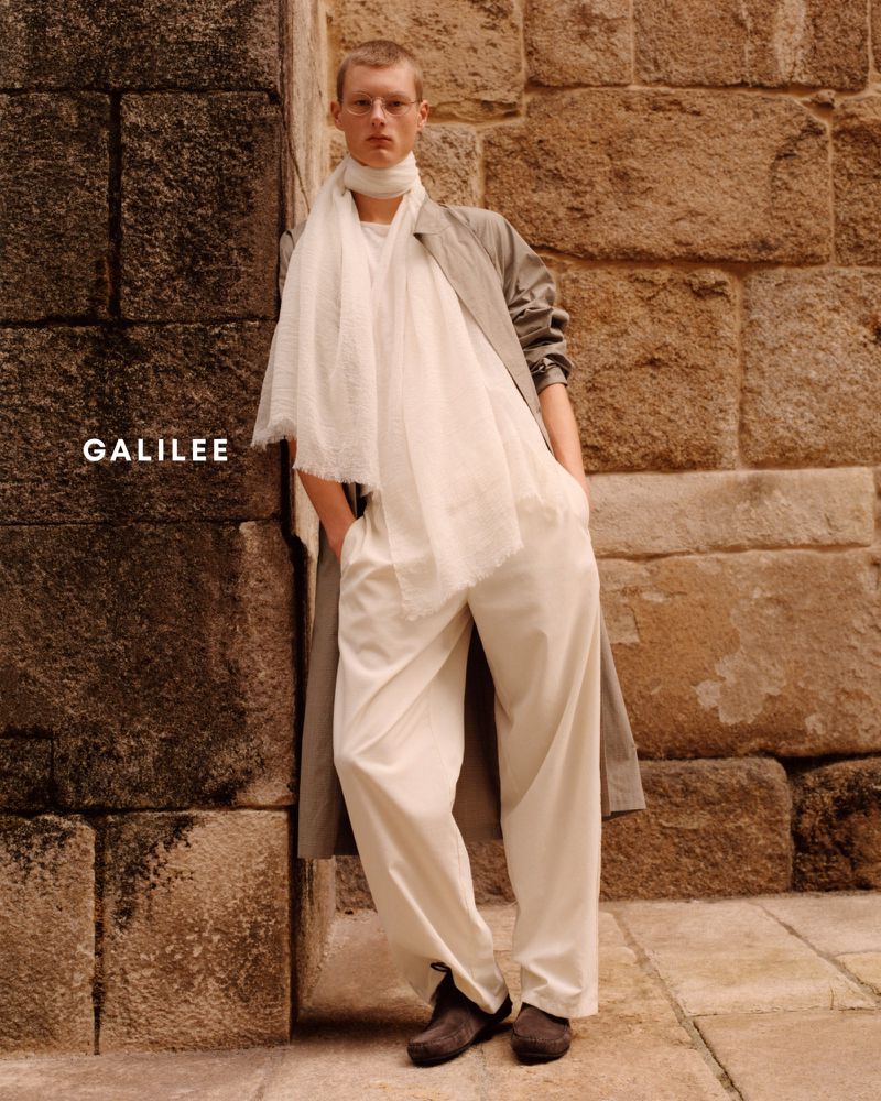 A male model wearing a grey coat, white scarf, and cream trousers leaning against a weathered stone wall.