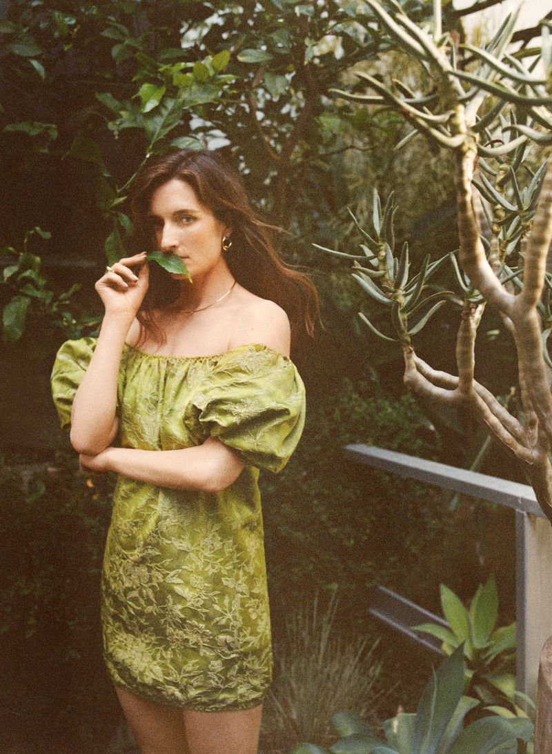 Young woman in a short, green off-the-shoulder dress poses with a leaf in an outdoor garden setting surrounded by green plants.