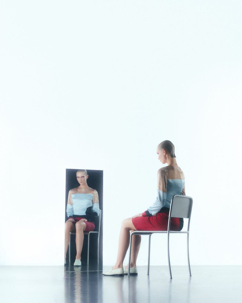 A female model seated on a metal chair looking into a vertical mirror reflection against a white background.