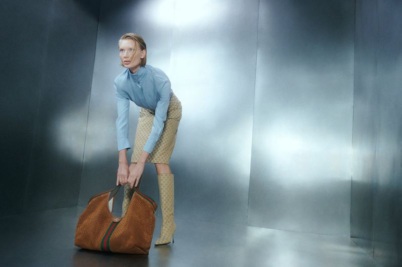 Model Yanita poses bending forward holding a large brown tote bag in a metallic studio space for L'Officiel Chile.