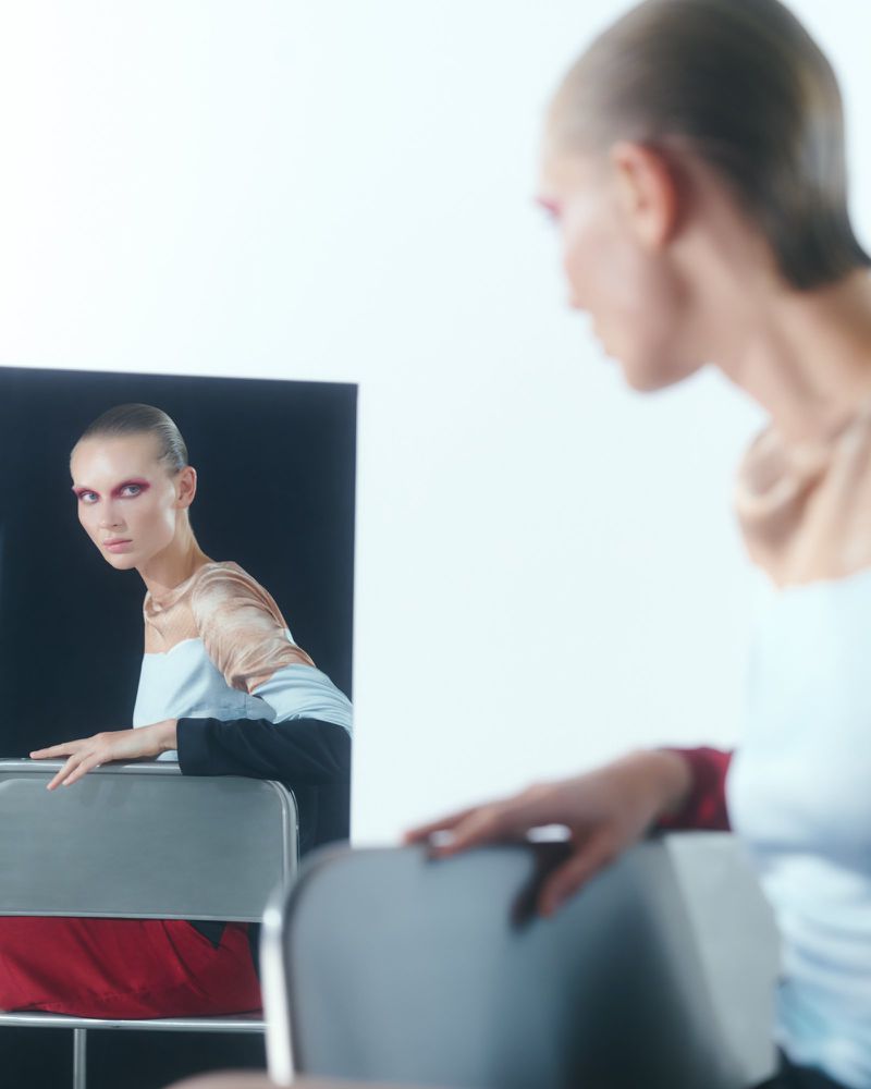 Fashion model reflected in a rectangular mirror wearing graphic red eye makeup and a color-blocked top against a bright white background.