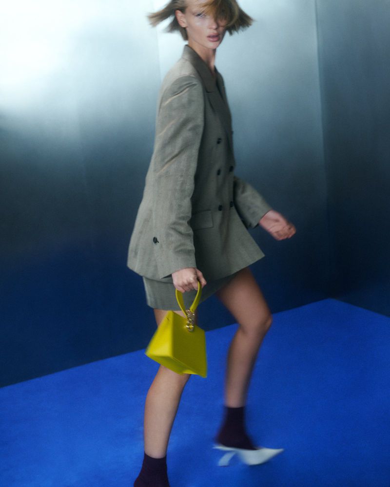 Model in grey blazer dress and white heels holding a yellow handbag against a blue floor and metallic gradient background