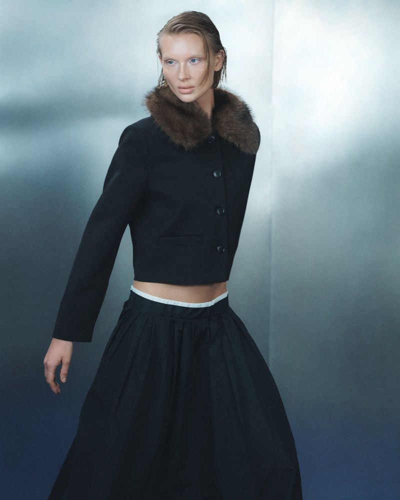 Model wearing a black cropped jacket with a brown fur collar and full black skirt against a cool gradient backdrop.