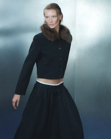 Model wearing a black cropped jacket with a brown fur collar and full black skirt against a cool gradient backdrop.