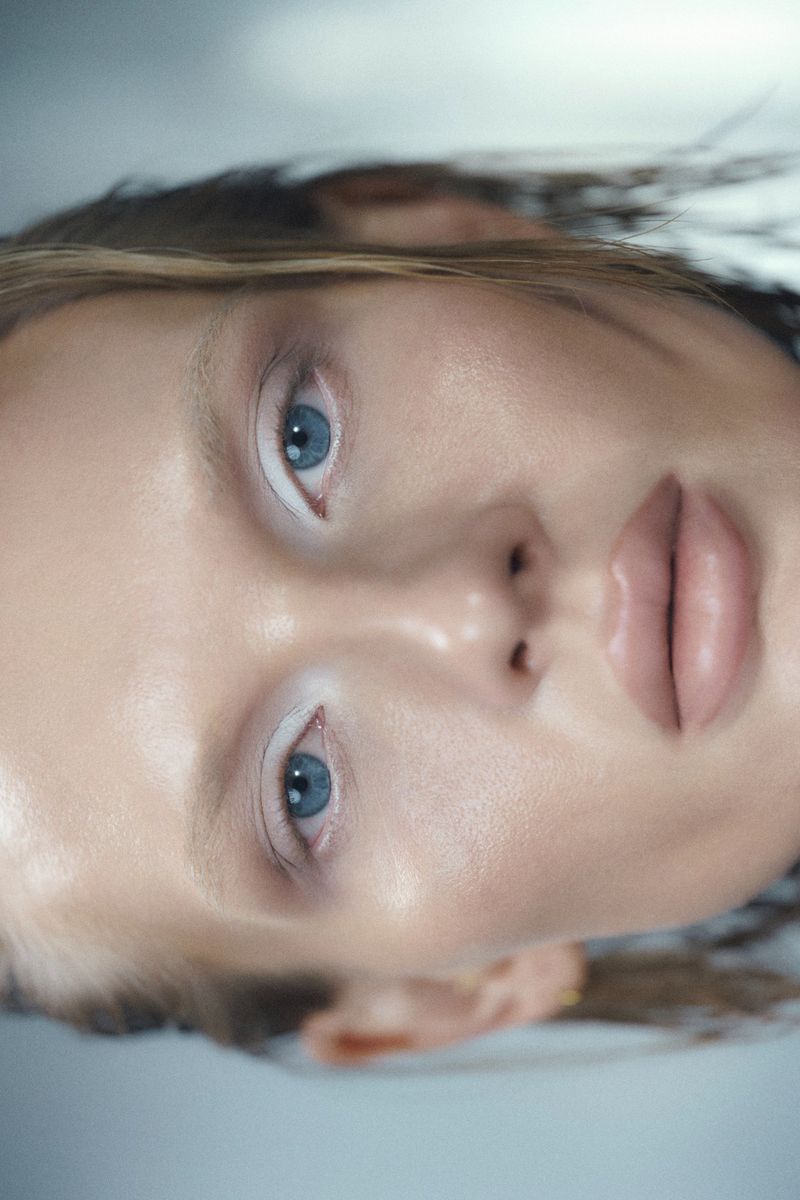 Close-up beauty portrait of a woman with white eyeliner, light bleached eyebrows, blue eyes, and slicked-back wet hair.