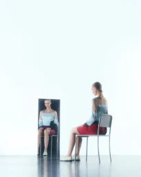 A female model seated on a metal chair looking into a vertical mirror reflection against a white background.