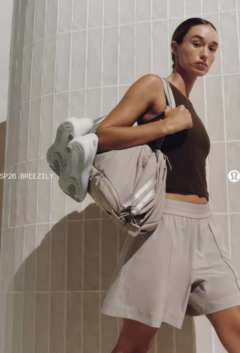 Model carrying a light grey duffel bag and white sneakers against a curved tile wall.