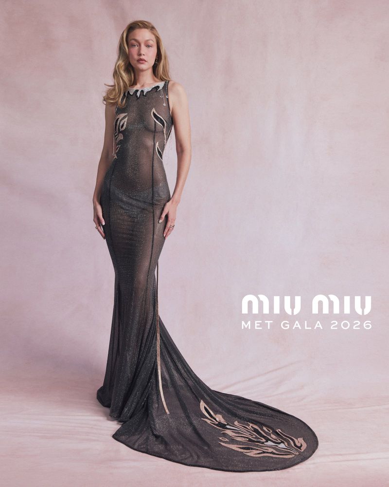 Gigi Hadid posing in a sheer charcoal embellished Miu Miu gown with an extended train against a soft pink backdrop.