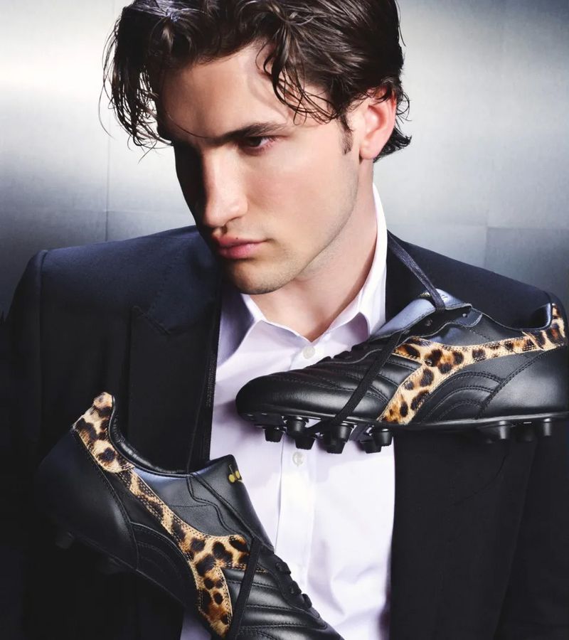 Portrait of adult man wearing black suit jacket, holding pair of black soccer shoes with leopard print design on grey background.