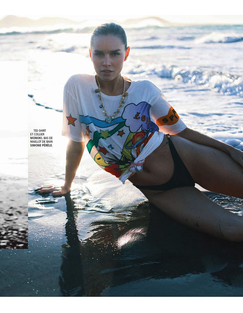 Olivia Aarnio reclines on wet dark sand wearing a white graphic t-shirt and black bikini bottoms beside ocean waves.