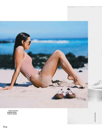 Model reclining in a red and white striped swimsuit and sunglasses on a sandy beach beside the blue ocean.