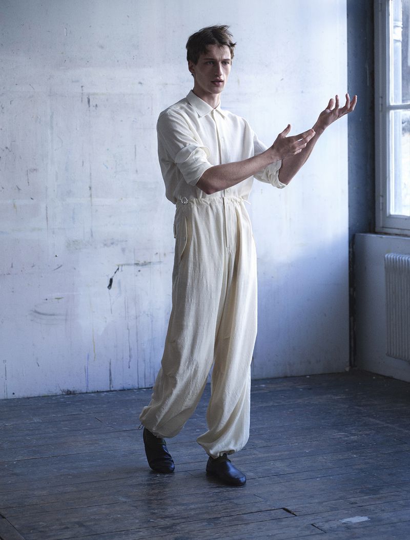 Model Vasko Luyckx posing in an off-white jumpsuit and dark shoes on dark wood flooring in front of a textured wall.