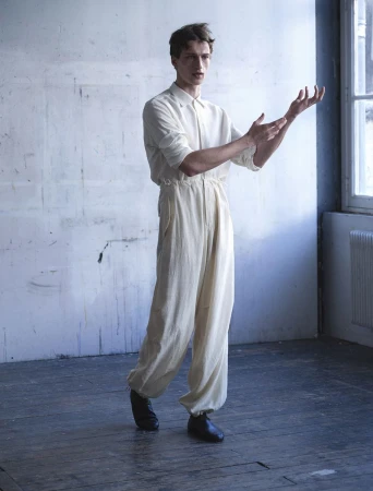Model Vasko Luyckx posing in an off-white jumpsuit and dark shoes on dark wood flooring in front of a textured wall.