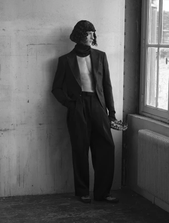 Model Vasko Luyckx wearing an unbuttoned black suit with bare chest holding a crown by a window.