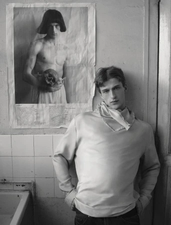 Model Vasko Luyckx posing in a light cowl-neck sweatshirt against a wall beside a monochrome poster of a man holding a skull.