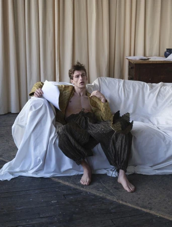 Model Vasko Luyckx wearing an open brocade jacket and voluminous gathered trousers seated on a draped white sofa holding a script.