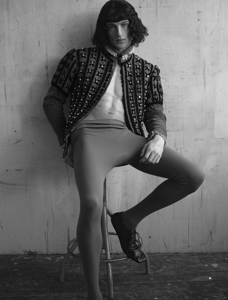 Model Vasko Luyckx seated on a stool wearing an open patterned doublet and tights in a monochrome studio portrait.