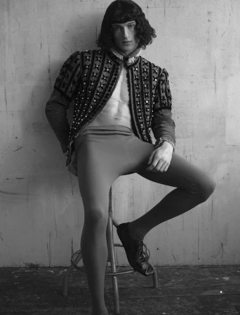 Model Vasko Luyckx seated on a stool wearing an open patterned doublet and tights in a monochrome studio portrait.