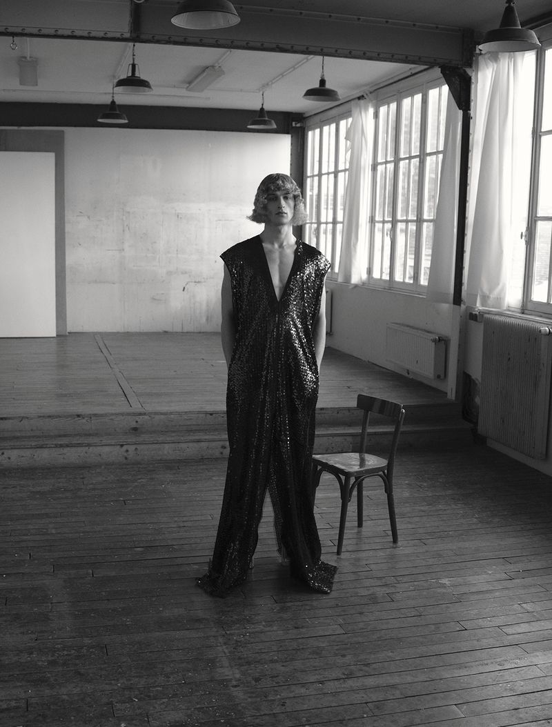 Model Vasko Luyckx wearing a black sequined sleeveless jumpsuit standing in an airy studio beside a wooden chair.