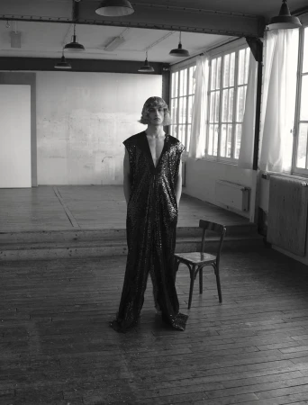 Model Vasko Luyckx wearing a black sequined sleeveless jumpsuit standing in an airy studio beside a wooden chair.