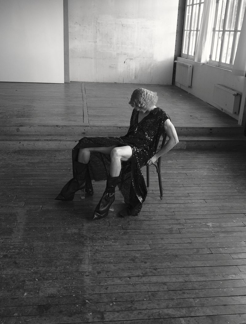 Model wearing a dark sequined dress and black boots seated on a wooden chair on distressed wood flooring.