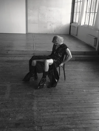 Model wearing a dark sequined dress and black boots seated on a wooden chair on distressed wood flooring.