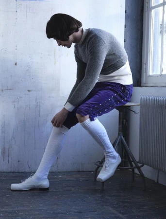 Model seated on a stool wearing a grey sweater, purple knee-length shorts, white socks, and white shoes beside a window.
