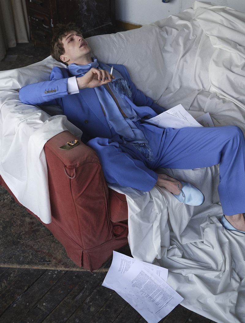 Young man in a blue suit reclining on a white-draped sofa holding a cigarette