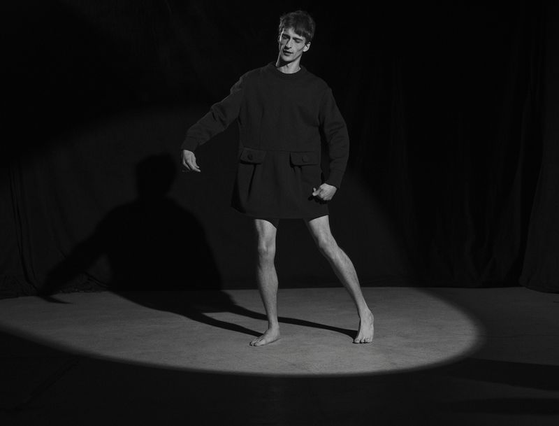 Barefoot male model in a black structured tunic posing within an oval spotlight beam on a dark stage in black and white.