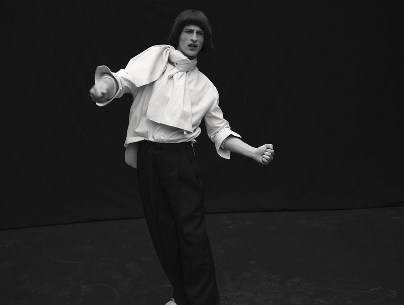 Model Vasko Luyckx wearing a draped white shirt and black trousers striking a dynamic pose against a black studio backdrop.