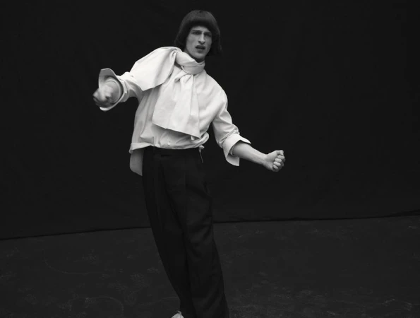 Model Vasko Luyckx wearing a draped white shirt and black trousers striking a dynamic pose against a black studio backdrop.