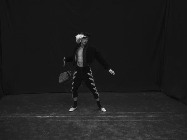 Model Vasko Luyckx posing in a feathered hat, open cropped jacket, and embellished breeches against a black studio backdrop.