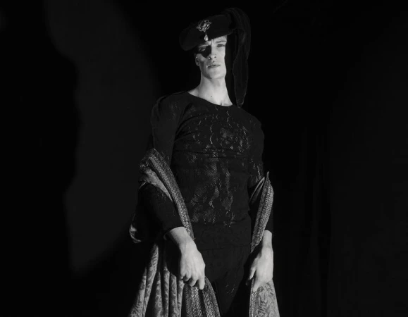 Model Vasko Luyckx posed in a black lace top, Renaissance cap with a pearl brooch, and draped patterned shawl on a dark background.