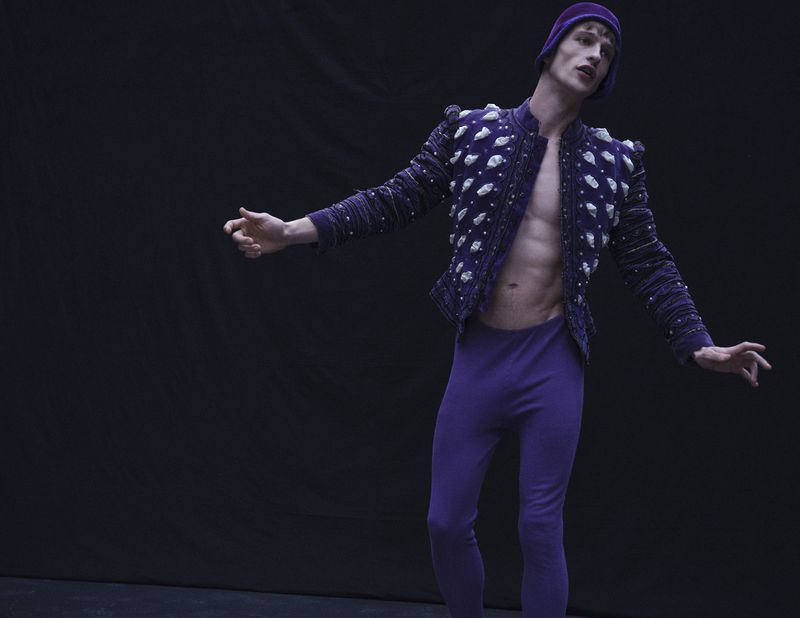 Model Vasko Luyckx poses in an open purple embellished doublet, matching purple tights, and a cap against a dark black background.