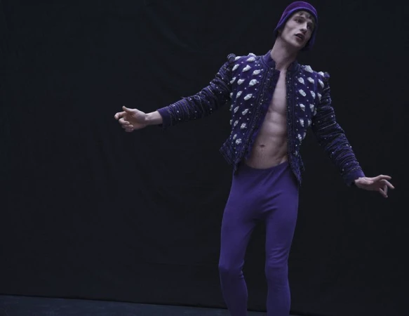 Model Vasko Luyckx poses in an open purple embellished doublet, matching purple tights, and a cap against a dark black background.