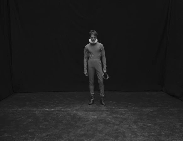 Male model Vasko Luyckx wearing a fitted grey outfit with a white neck ruff and holding a wreath against a dark backdrop.