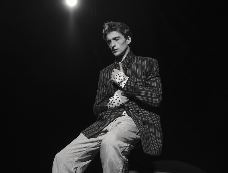Male model seated in a dark pinstripe jacket and polka dot shirt under dramatic spotlight against a black background.